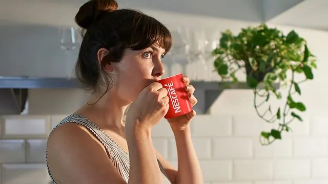 Woman with her hair in a bun drinking from a red NESCAFÉ mug in a kitchen with a plant on a shelf behind her, the image used on the Smooth Radio NESCAFÉ competition page. It does not show the prize.