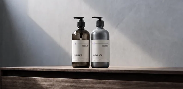Two WØRKS botanical hand and body wash bottles, one amber and one grey, standing on a wooden shelf against a grey wall.